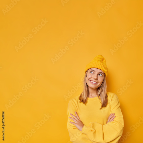 Pleased young cute woman with dreamy expression keeps arms folded stands in assertive pose wears hat and casual jumper isolated over yellow background copy space area for your advertisement.