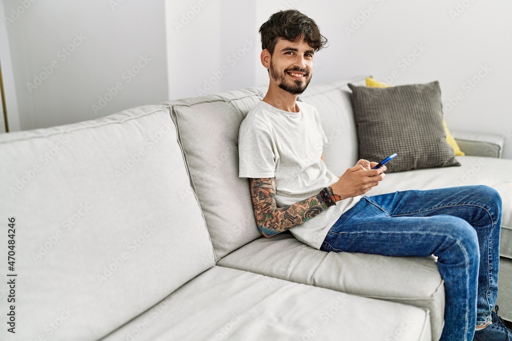 Young hispanic man using smartphone sitting on the sofa at home.