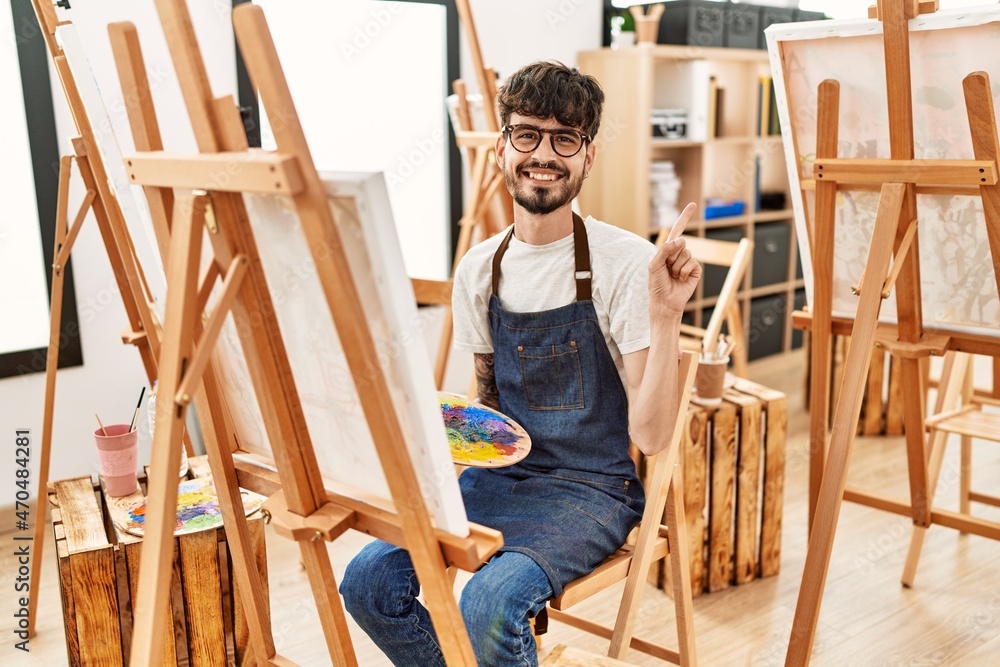 Hispanic man with beard at art studio smiling happy pointing with hand and finger to the side Hispanic man with beard at art studio smiling happy pointing with hand and finger to the side