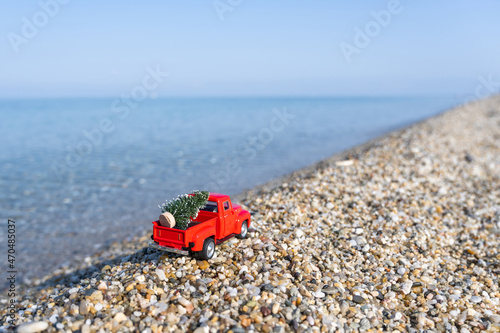 Red retro small car carrying christmas tree on the sea beach shore. Winter holiday concept