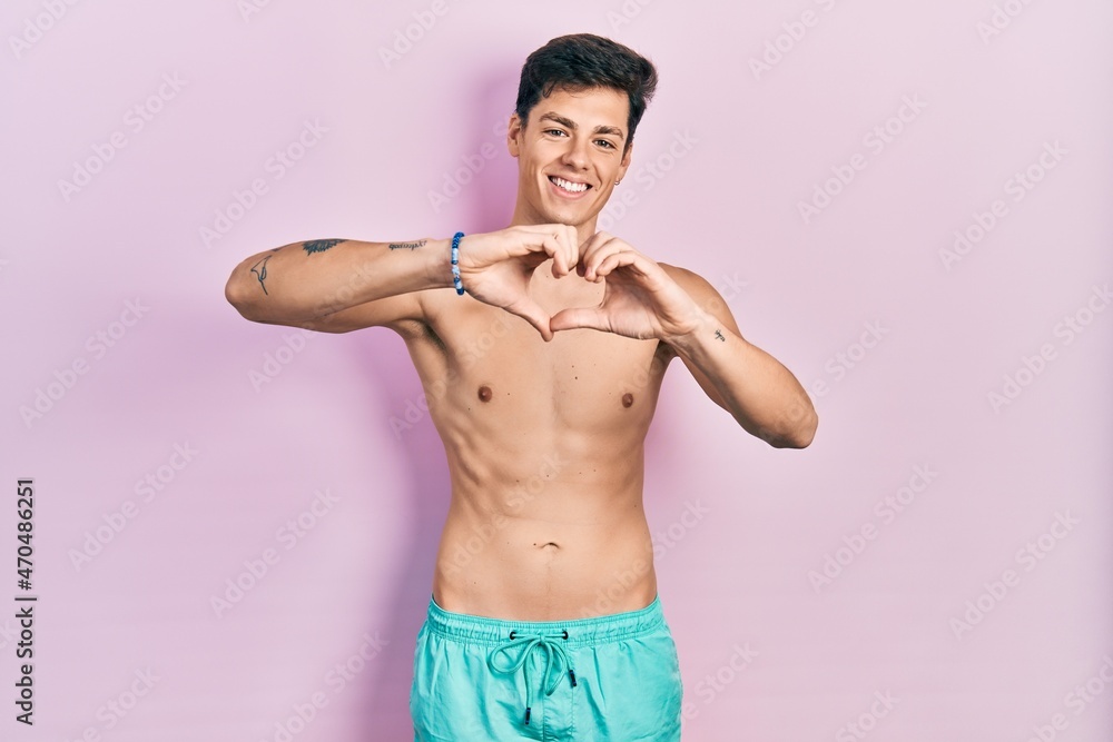 Young hispanic man wearing swimwear shirtless smiling in love doing heart symbol shape with hands. romantic concept.