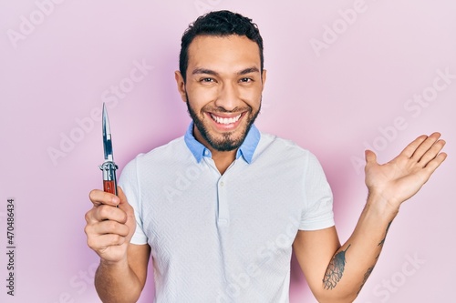 Hispanic man with beard holding pocket knife celebrating victory with happy smile and winner expression with raised hands