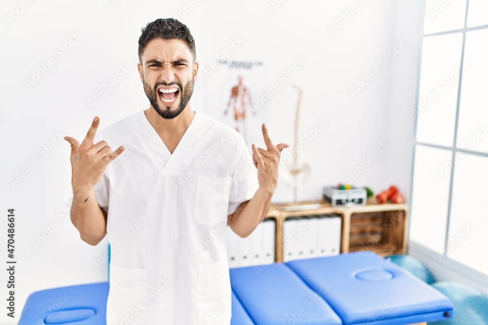 Young handsome man with beard working at pain recovery clinic shouting with crazy expression doing rock symbol with hands up. music star. heavy concept. Young handsome man with beard working at pain recovery clinic shouting with crazy expression doing rock symbol with hands up. music star. heavy concept.