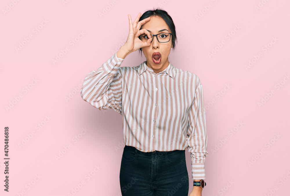 Fototapeta premium Young hispanic girl wearing casual clothes and glasses doing ok gesture shocked with surprised face, eye looking through fingers. unbelieving expression.