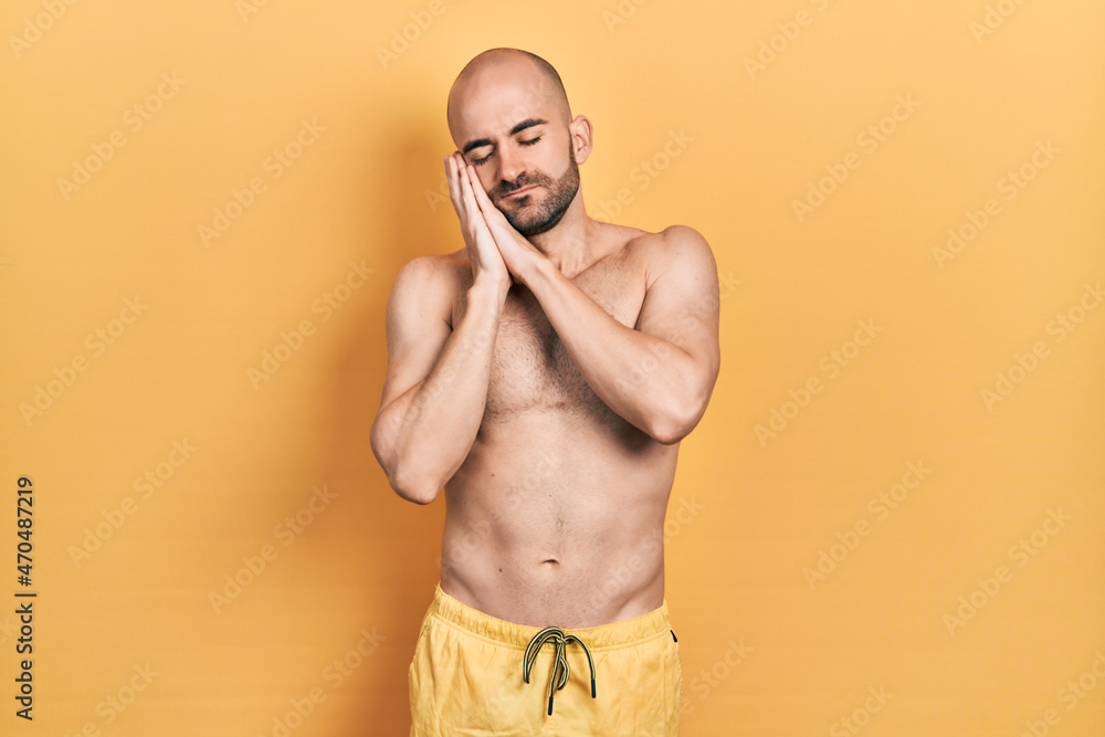 Young bald man wearing swimwear sleeping tired dreaming and posing with hands together while smiling with closed eyes.