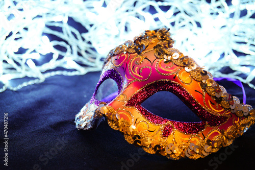 Purple and yellow carnival mask on black silk. There are festive white lights in the background.