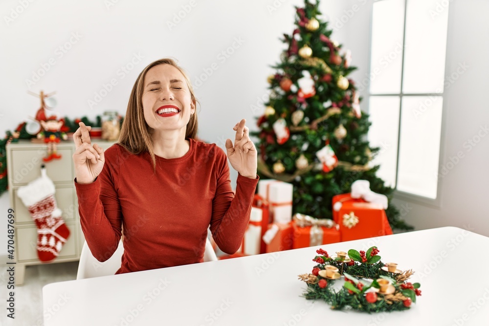 Caucasian young blonde woman sitting on the table by christmas tree gesturing finger crossed smiling with hope and eyes closed. luck and superstitious concept. Caucasian young blonde woman sitting on the table by christmas tree gesturing finger crossed smiling with hope and eyes closed. luck and superstitious concept.