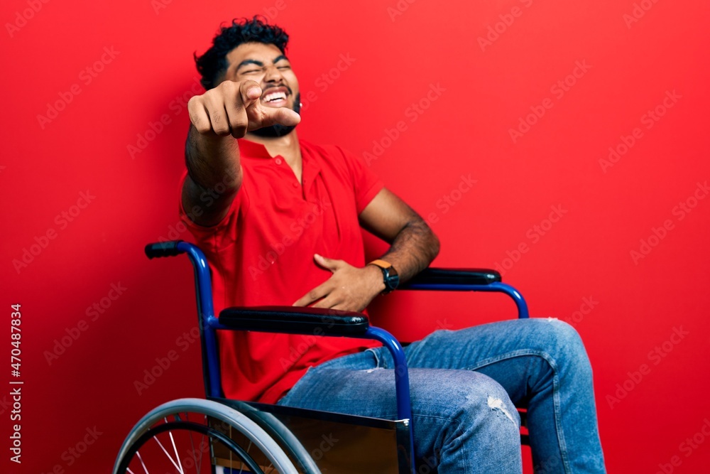 Arab man with beard sitting on wheelchair laughing at you, pointing finger to the camera with hand over body, shame expression