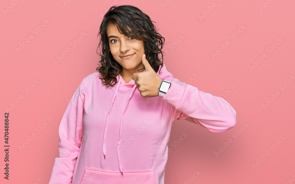 Young hispanic woman wearing casual sweatshirt doing happy thumbs up gesture with hand. approving expression looking at the camera showing success. Young hispanic woman wearing casual sweatshirt doing happy thumbs up gesture with hand. approving expression looking at the camera showing success.