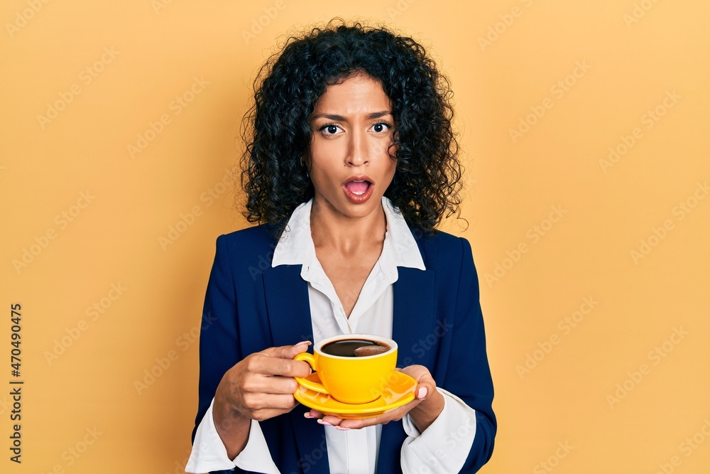 Young latin girl wearing business style drinking cup of coffee in shock face, looking skeptical and sarcastic, surprised with open mouth Young latin girl wearing business style drinking cup of coffee in shock face, looking skeptical and sarcastic, surprised with open mouth