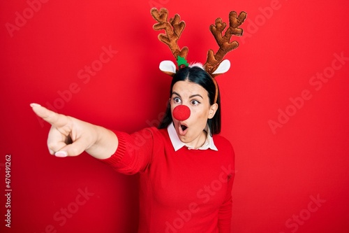 Young hispanic woman wearing deer christmas hat and red nose pointing with finger surprised ahead, open mouth amazed expression, something on the front