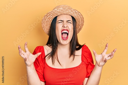 Young hispanic woman wearing summer hat crazy and mad shouting and yelling with aggressive expression and arms raised. frustration concept.