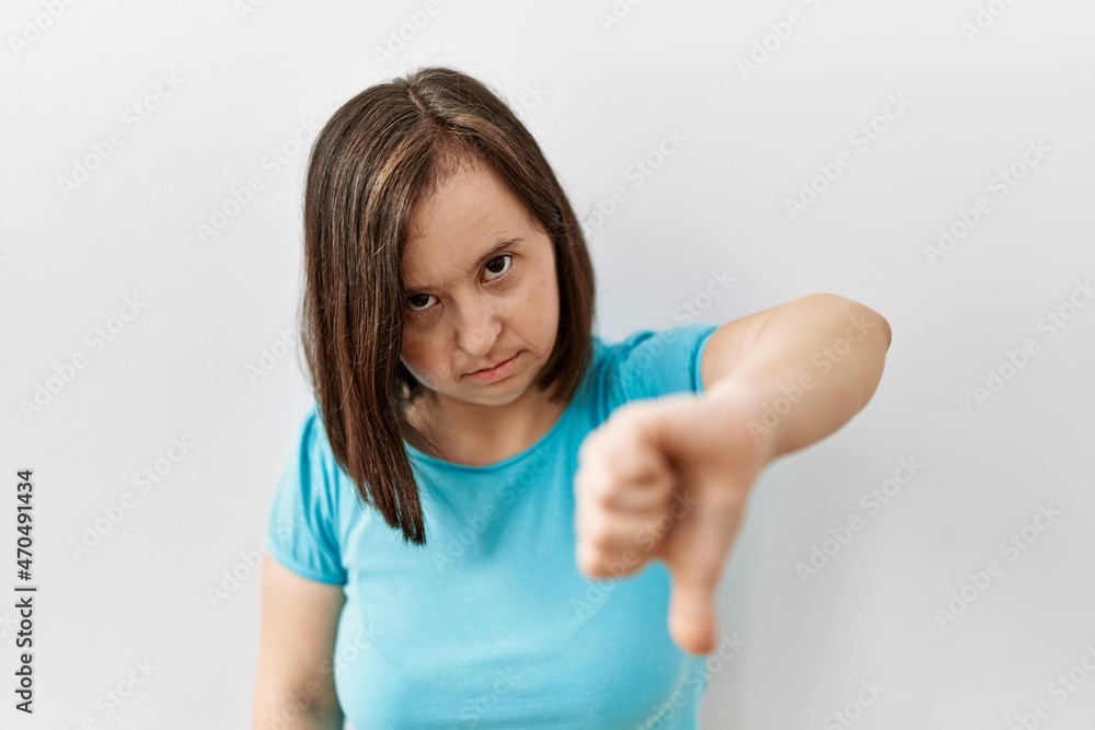 Young down syndrome woman standing over isolated background looking unhappy and angry showing rejection and negative with thumbs down gesture. bad expression. Young down syndrome woman standing over isolated background looking unhappy and angry showing rejection and negative with thumbs down gesture. bad expression.