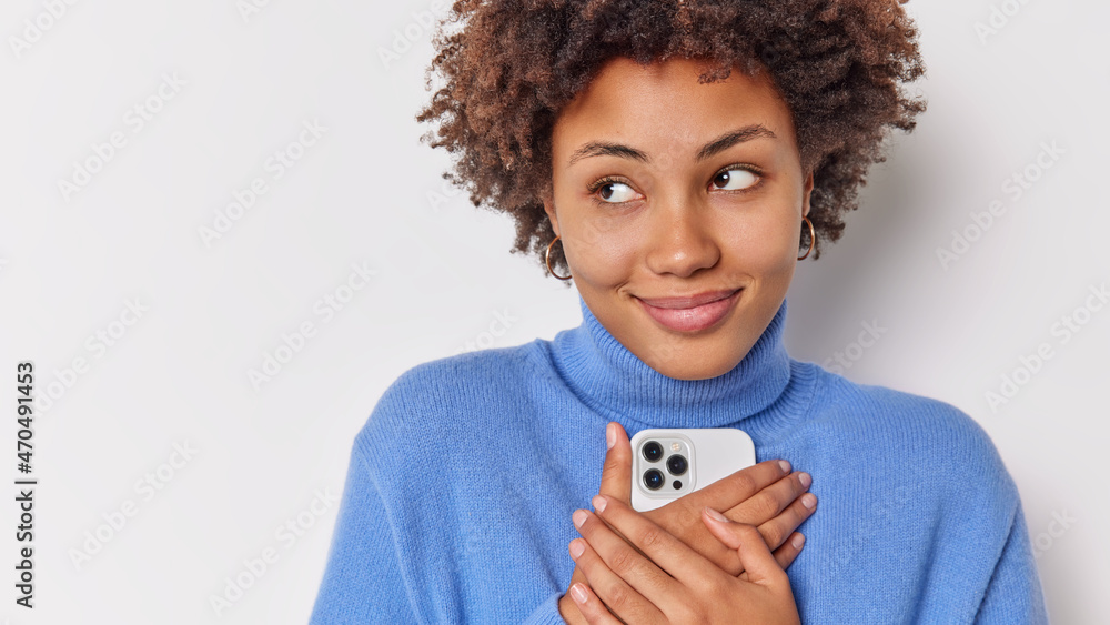 Photo of lovely curly haired woman concentrated aside pensively has dreamy expression holds mobile phone closely to body glad to receive message from boyfriend isolated over white background Photo of lovely curly haired woman concentrated aside pensively has dreamy expression holds mobile phone closely to body glad to receive message from boyfriend isolated over white background