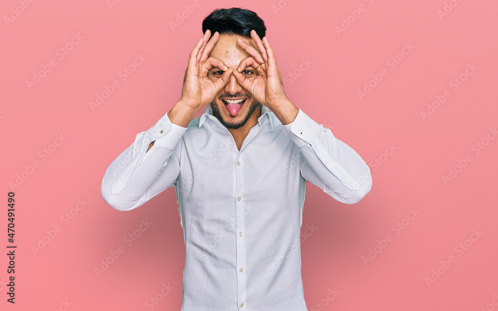 Young arab man wearing casual clothes doing ok gesture like binoculars sticking tongue out, eyes looking through fingers. crazy expression. Young arab man wearing casual clothes doing ok gesture like binoculars sticking tongue out, eyes looking through fingers. crazy expression.