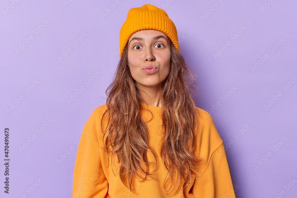 Photo of romantic young woman with folded lips looks surprisingly at camera has flirty expression wears casual jumper and hat isolated over purple background blows air kiss makes smack gesture Photo of romantic young woman with folded lips looks surprisingly at camera has flirty expression wears casual jumper and hat isolated over purple background blows air kiss makes smack gesture