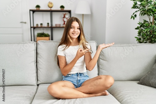 Young brunette teenager sitting on the sofa at home amazed and smiling to the camera while presenting with hand and pointing with finger.