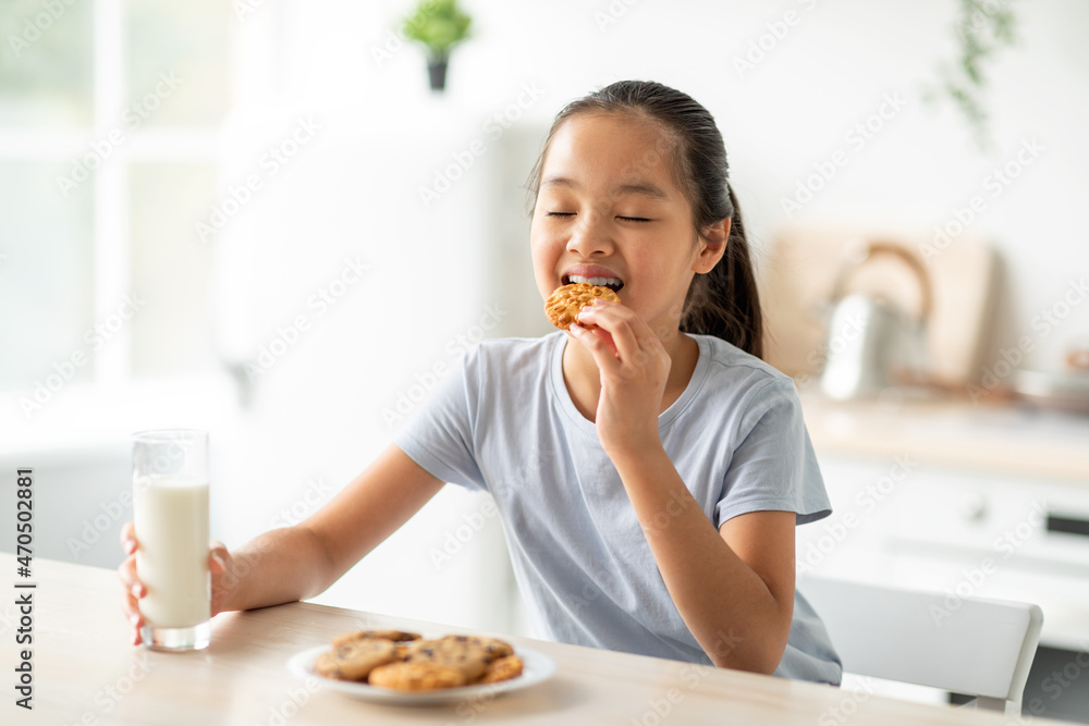Pretty asian girl eating cookies and drinking milk in kitchen, happy female child enjoying healthy snack at home Pretty asian girl eating cookies and drinking milk in kitchen, happy female child enjoying healthy snack at home