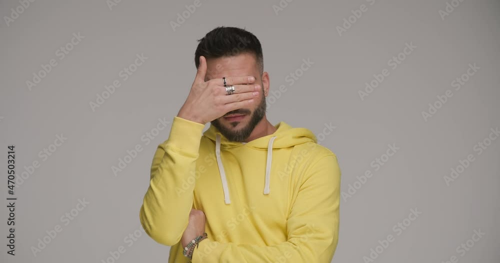 sexy bearded man in yellow sweatshirt playing hide and seek with hand, making funny faces, looking to side on grey background sexy bearded man in yellow sweatshirt playing hide and seek with hand, making funny faces, looking to side on grey background