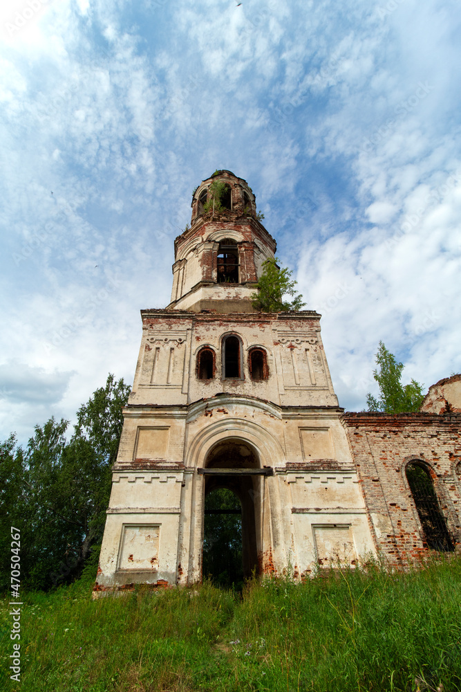View of the front side of the bell tower of an old, destroyed and abandoned church in Russia. Walls with peeling paint and old red bricks. Trees on the roofs. Summer. Daylight. Sky with clouds.