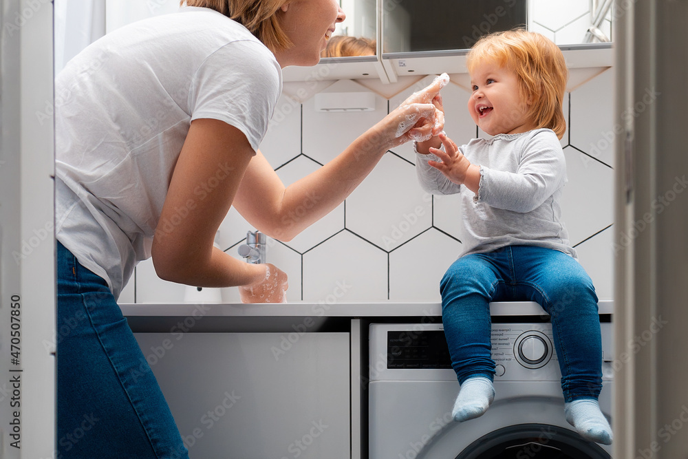 A young happy mother with foam on her hands is playing with her daughter. A little child learns from his mother how to wash hands. Hygienic procedures A young happy mother with foam on her hands is playing with her daughter. A little child learns from his mother how to wash hands. Hygienic procedures