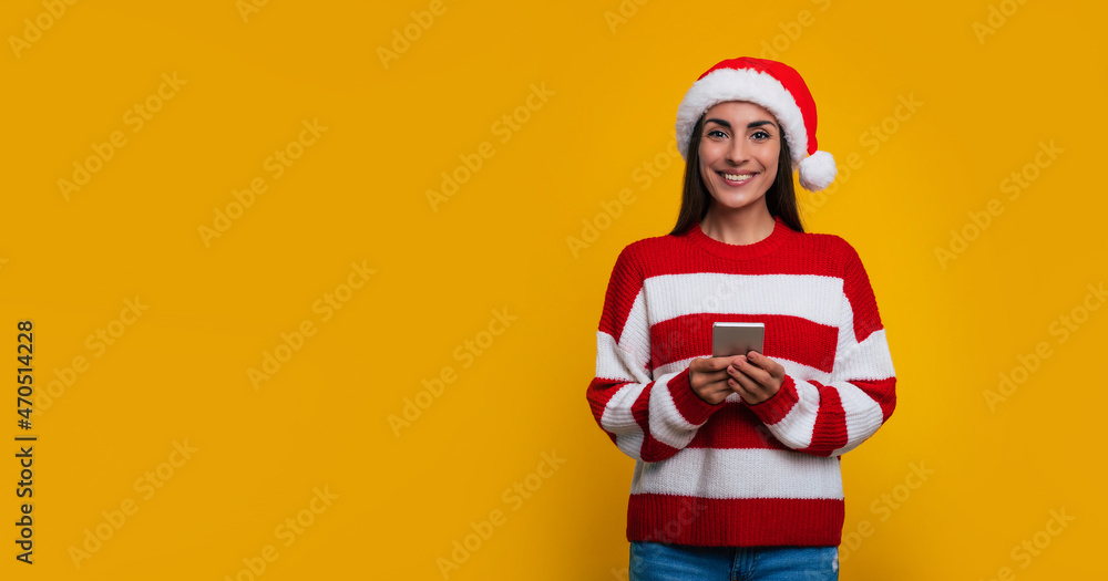Banner photo of happy beautiful woman in Santa hat with smart phone in hands using it and looking with smile on camera Banner photo of happy beautiful woman in Santa hat with smart phone in hands using it and looking with smile on camera
