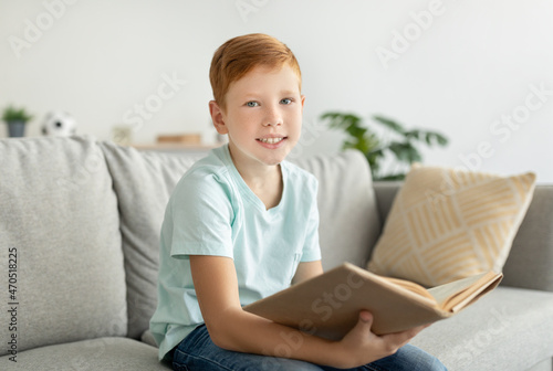 Joyful ginger boy sitting on couch at home, reading book