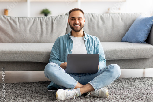 Full length of happy young guy sitting cross legged on floor, using laptop computer, smiling at camera indoors
