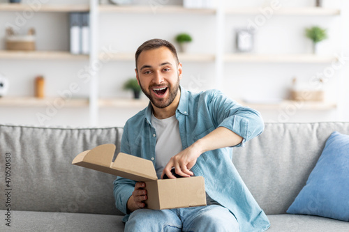 Portrait of cheerful young man receiving parcel, unboxing package, taking out gaming joystick, sitting on couch at home