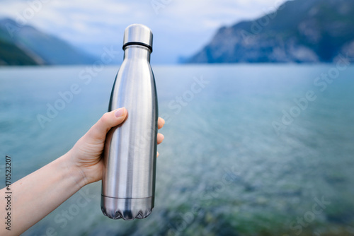 Close-up of female hand holding reusable, steel thermo shiny bottle for water, on the background of clear water of a lake with a turquoise hue. Copy space concept.