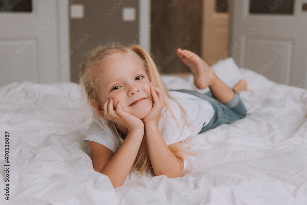 portrait of little smiling blonde girl in a white T-shirt and jeans lying on her stomach in bed with white bedding. children's recreation. space for text. High quality photo portrait of little smiling blonde girl in a white T-shirt and jeans lying on her stomach in bed with white bedding. children's recreation. space for text. High quality photo