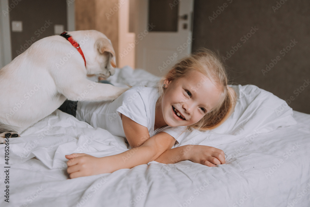 portrait of a little blonde girl lying in bed with her beloved dog in her arms. girl in a white T-shirt and jeans and a pet are lying on white bed linen. space for text. High quality photo portrait of a little blonde girl lying in bed with her beloved dog in her arms. girl in a white T-shirt and jeans and a pet are lying on white bed linen. space for text. High quality photo