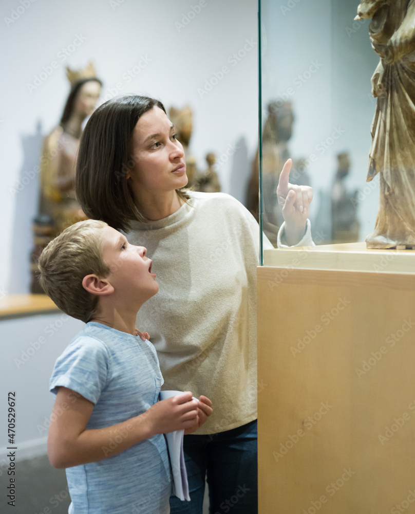 Young woman with son observing with interest sculptures exhibition in art museum, pointing to something interesting
