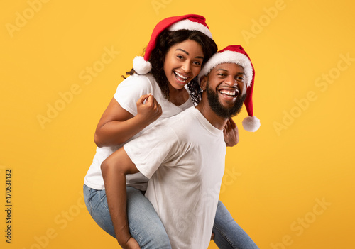 Happy black guy giving his girlfriend piggyback ride, wearing together Santa hats, having fun over yellow background © Prostock-studio