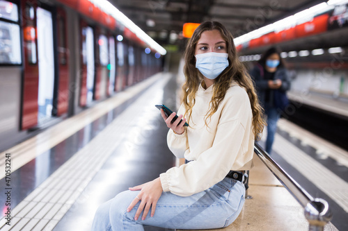 Girl wearing a protective mask, sitting on a subway platform bench during a pandemic, texts with friends on her mobile ..phone while waiting for a train