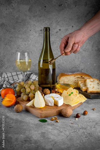 A board with cheese, a bottle of wine and fruit on a gray background.
A man's hand pours honey on top of camembert cheese