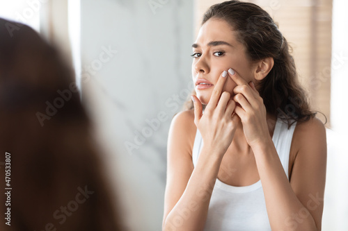 Unhappy Female Squeezing Pimples On Cheek Looking At Mirror Indoor