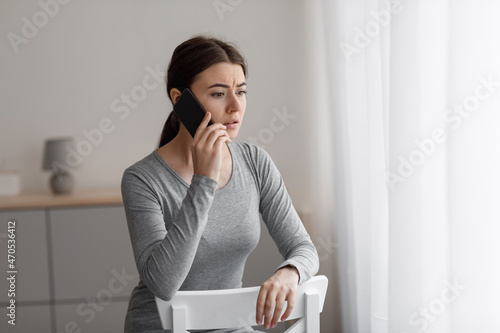 Sad unhappy worried european millennial female sitting on chair and talking on phone at home photo
