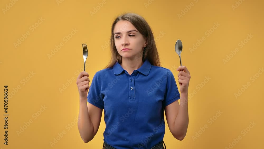 Portrait sad woman on diet choosing between fork and spoon suffering hungry isolated on orange Portrait sad woman on diet choosing between fork and spoon suffering hungry isolated on orange