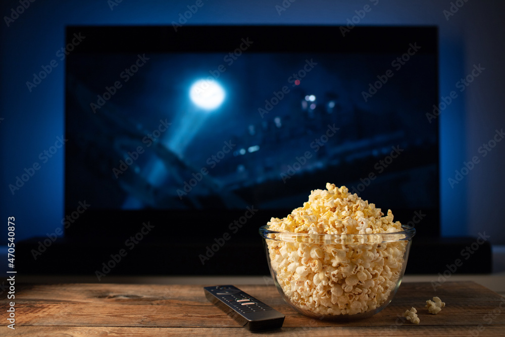 A glass bowl of popcorn and remote control in the background the TV works.  Evening cozy watching a movie or TV series at home Photos | Adobe Stock