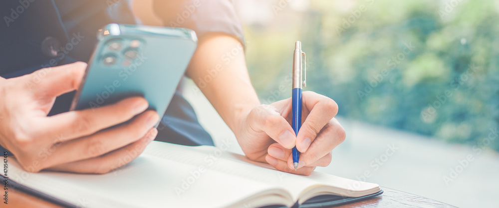 Woman hand writing on a notepad with a pen and she is holding a cell phone in the office.