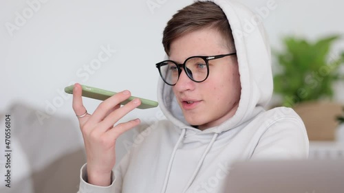 Online education of teen students at home. Schoolboy in white hoodie spbi with glasses records audio message on smartphone near laptop computer at table closeup 4k video photo