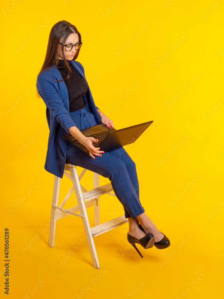 Stylish businesswoman with laptop. Woman is dressed in modern business attire. Attractive girl with computer. She is sitting in high chair. Lady concept using business software. Girl laptop on yellow Stylish businesswoman with laptop. Woman is dressed in modern business attire. Attractive girl with computer. She is sitting in high chair. Lady concept using business software. Girl laptop on yellow