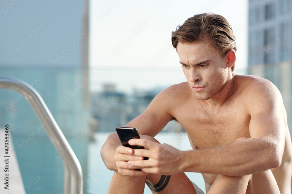 Frowning young man answering mean text messages on smartphone after swimming in outdoor pool Frowning young man answering mean text messages on smartphone after swimming in outdoor pool