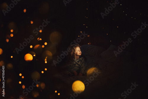 Fototapeta Naklejka Na Ścianę i Meble -  Christmas night, a child, a girl sitting in the dark with her eyes closed, dreaming, around Christmas lights, garlands.