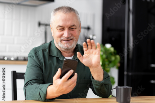 Elderly man communicates online using a mobile phone while sitting at home in the kitchen. Senior man holding smartphone in hands talking on video call