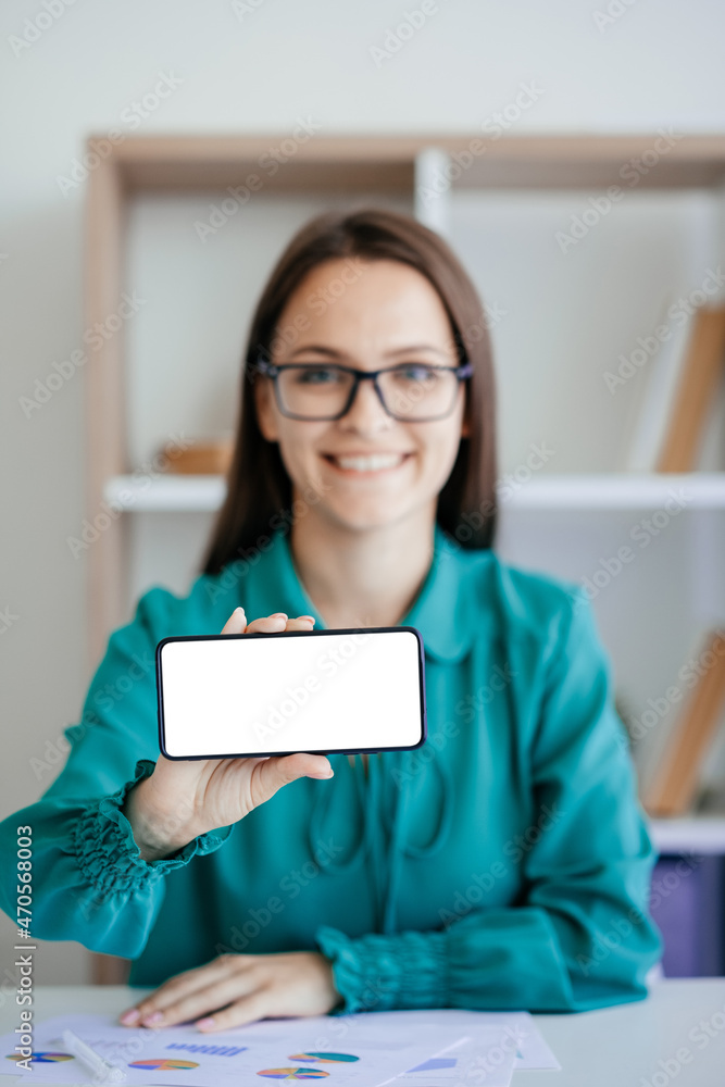 Digital marketing. Mobile business. Online professional communication. Smiling woman showing horizontal white empty mockup smartphone screen on defocused office background. Digital marketing. Mobile business. Online professional communication. Smiling woman showing horizontal white empty mockup smartphone screen on defocused office background.