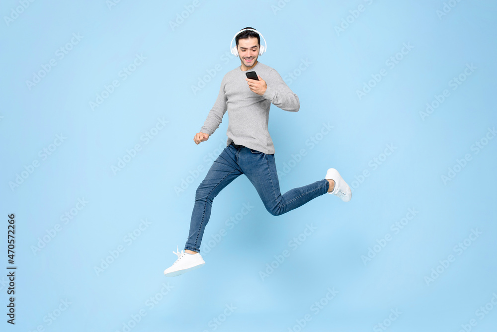 Energetic young Caucasian man wearing headphones using smartphone and jumping in studio light blue color isolated background Energetic young Caucasian man wearing headphones using smartphone and jumping in studio light blue color isolated background
