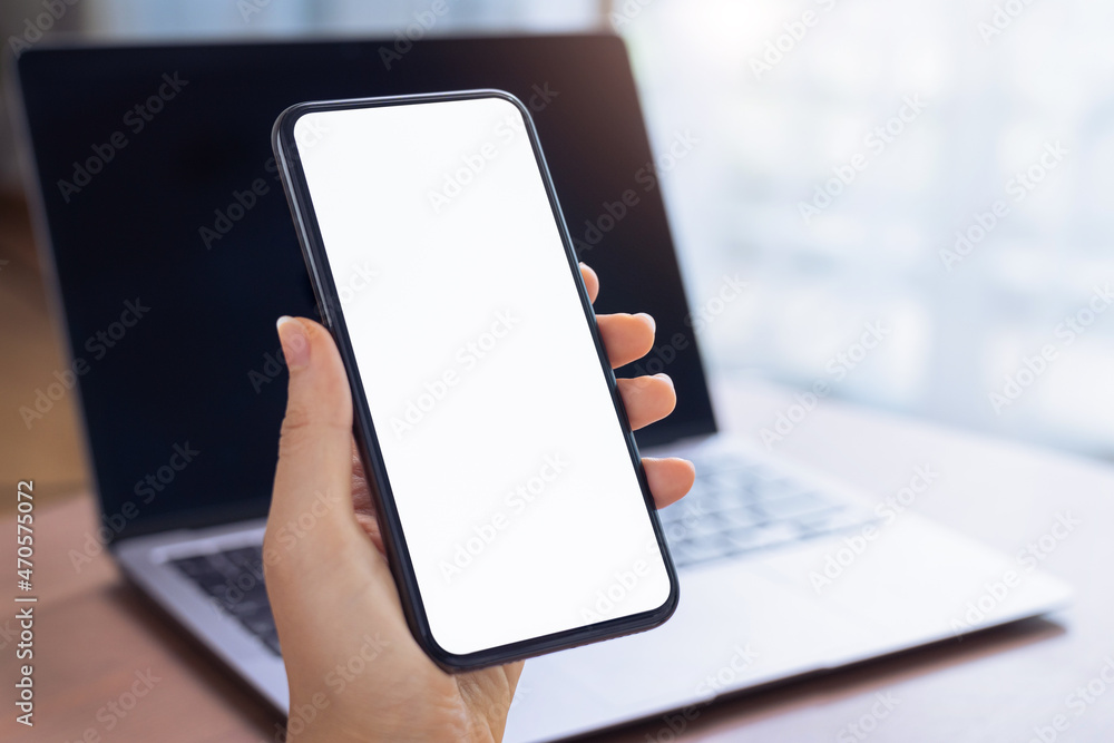 Close up of hand holding smartphone with white mock-up screen and laptop.. Close up of hand holding smartphone with white mock-up screen and laptop..