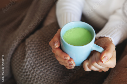 Close up of hands holding blue cup of matcha tea. Model in white sweater and cozy plaid is sitting at home. Depth of field and empty space on blurry beige background